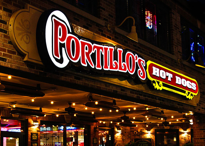Portillo's chain restaurant storefront at night with illuminated red and white signage warning about food orders.