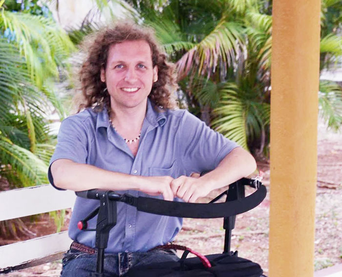 Man trapped in hospital for 9 years sitting outdoors, smiling and leaning on a walker with tropical plants in the background