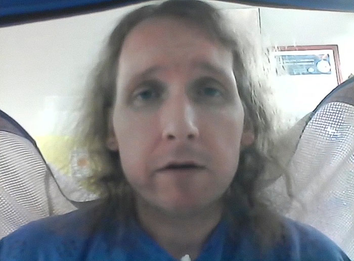 Man with long hair and blue shirt speaking on video call after being trapped in hospital for nine years exposing staff abuse.