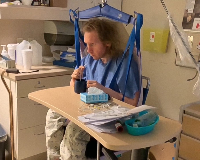Man trapped in hospital for 9 years sitting at a table in medical gown with hospital equipment around him.