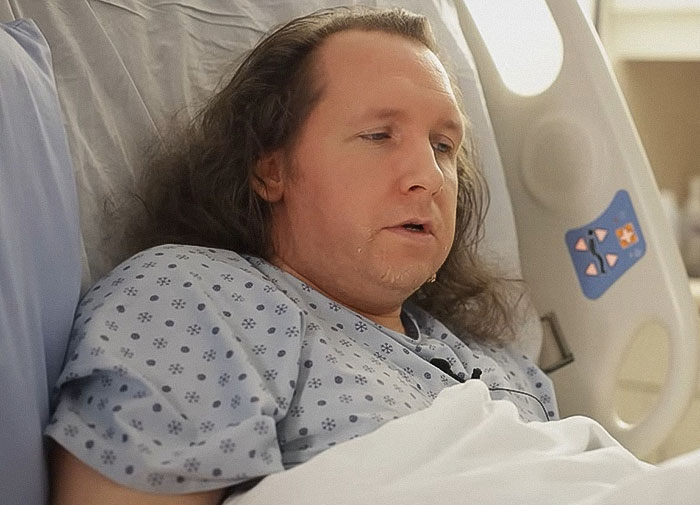 Man lying in hospital bed wearing patterned hospital gown, appearing distressed and fatigued during recovery.