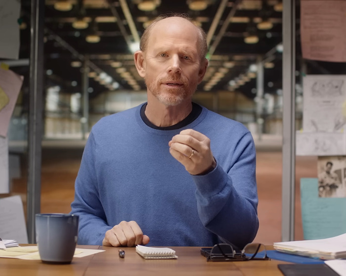 A man in a blue sweater seated at a desk with notes and a mug, discussing Ron Howard&rsquo;s Nepo Baby Bryce Dallas Howard.
