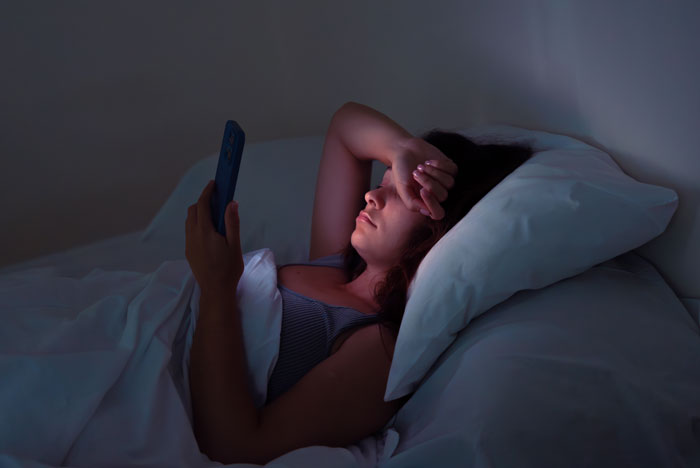 Woman in bed using smartphone at night, tired and restless during nightly gaming marathons, sleep deprivation evident.