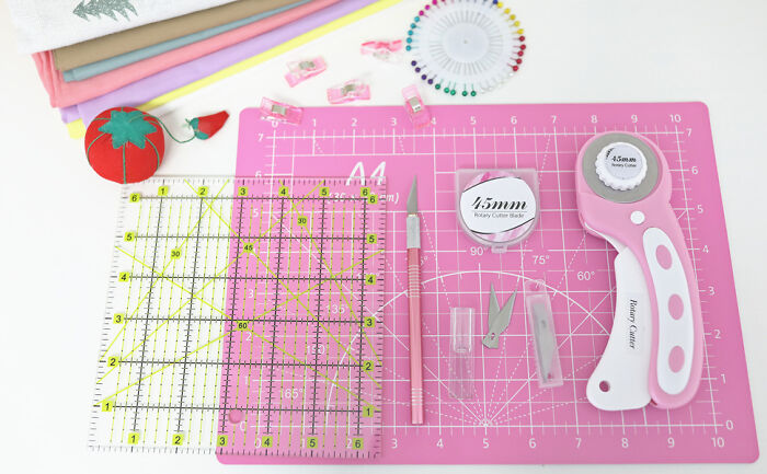 Pink crafting cutting mat with rotary cutter, ruler, craft knife, pins, and fabric pieces for sewing and crafting projects