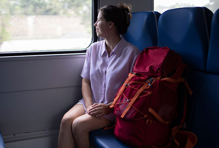 Young woman sitting on a train seat looking out the window with a large red backpack beside her during travel.