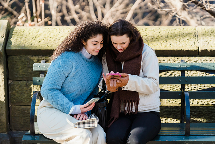 Teen niece and aunt sitting on park bench, niece showing phone and aunt holding wallet during a tense moment.