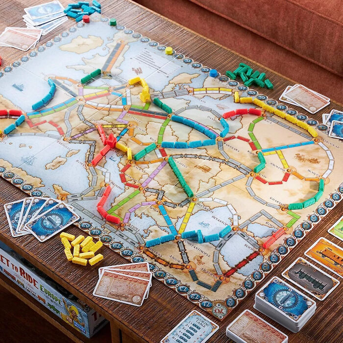 Board game setup of Ticket to Ride Europe with colorful train pieces and cards on a wooden table beside a couch.