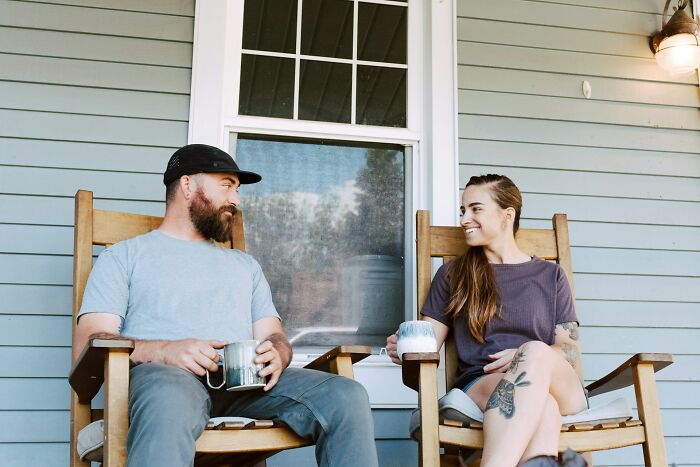 Couple sitting on wooden chairs outside, smiling and holding mugs, displaying weird things couples started doing together.
