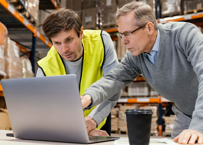Two men in a warehouse reviewing store management on a laptop, highlighting issues caused by a new manager's rule.