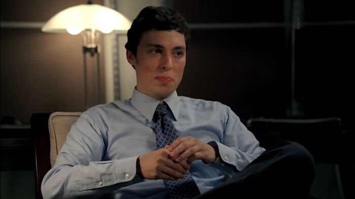 Young man in office attire sitting on a chair, representing tragic losses in TV show history in a dramatic scene.