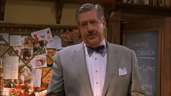 Man in a gray suit and bow tie standing indoors with a bulletin board and chalkboard in the background, depicting tragic losses in TV shows.