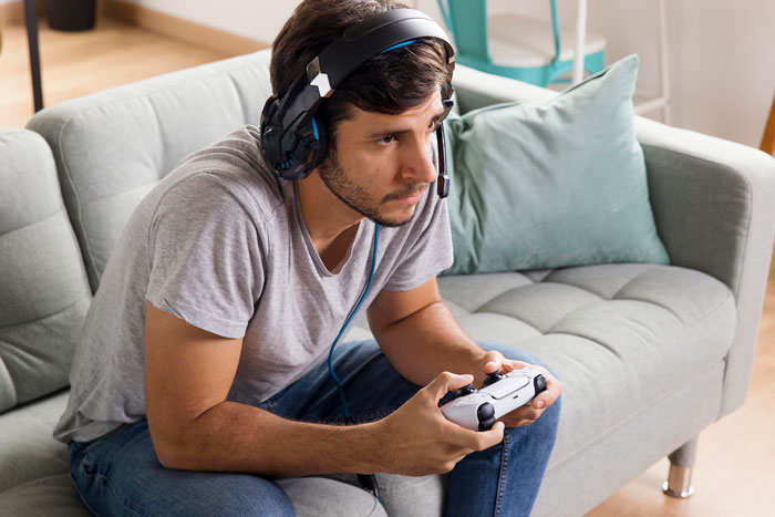 Young man with headset focused on exploratory testing while playing a game and reporting bugs on a gray couch. Young man with headset focused on exploratory testing while playing a game and reporting bugs on a gray couch.
