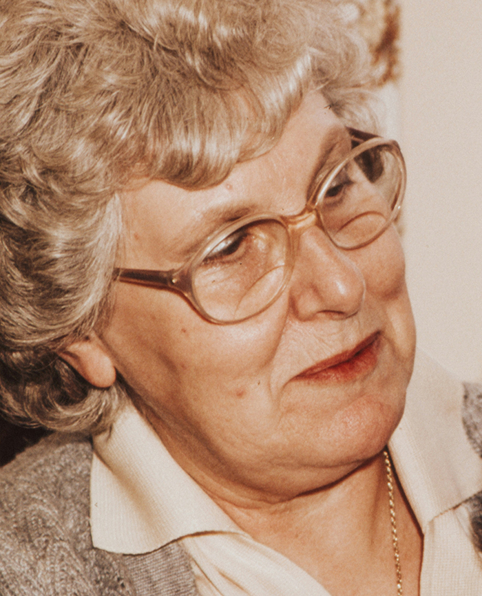 Elderly woman with glasses and gray hair, wearing a sweater, reflecting on disturbing deathbed confessions.