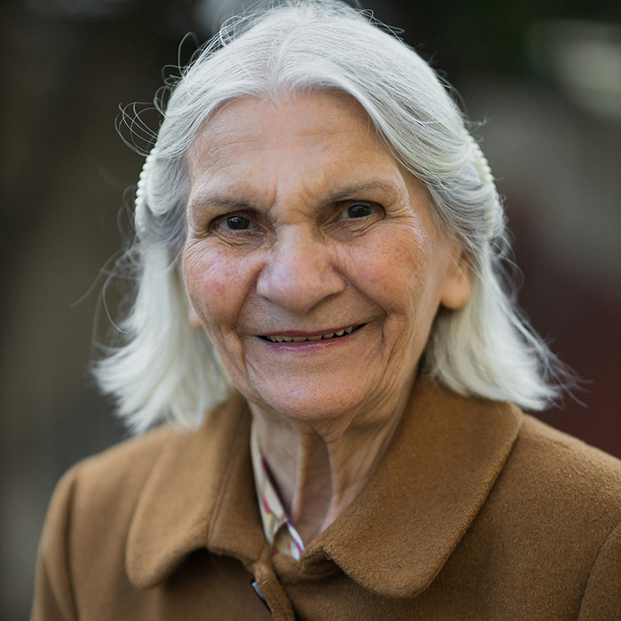 Elderly woman with white hair and a brown coat smiling gently, reflecting on disturbing deathbed confessions.