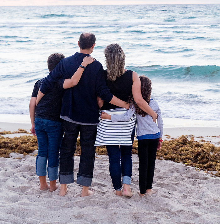 Family of four standing together on the beach, looking at the ocean, symbolizing reflection on deathbed confessions and guilt.