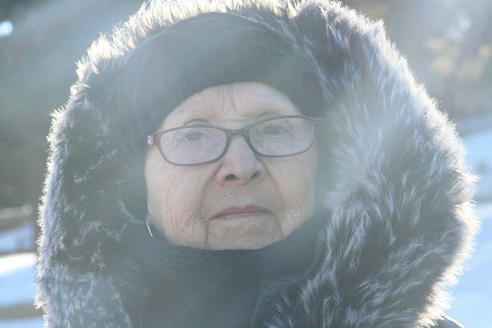 Elderly woman wearing glasses and a fur-lined hood, reflecting on disturbing deathbed confessions in a cold setting.