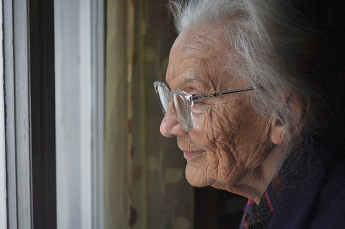 Elderly woman with glasses looking thoughtfully out a window, reflecting on disturbing deathbed confessions and guilt.