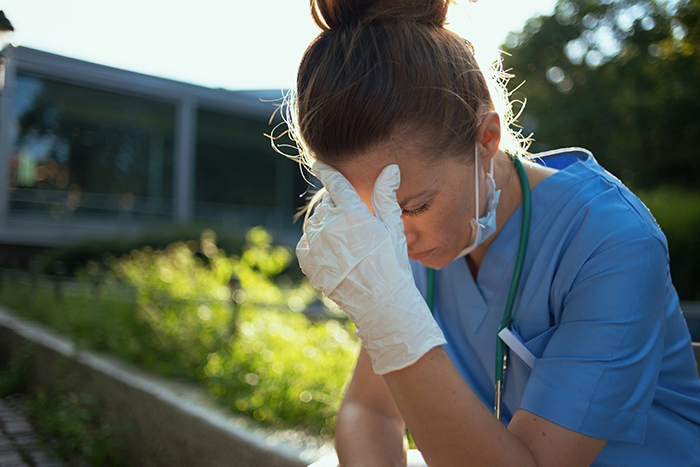 Stressed nurse in scrubs with gloves and stethoscope, holding her head outdoors, reflecting on disturbing deathbed confessions.