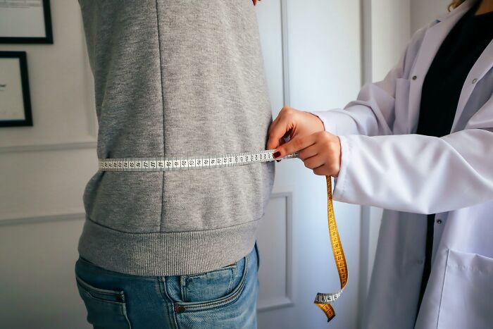 Person in grey sweatshirt having waist measured by healthcare professional using tape in emotional confession setting