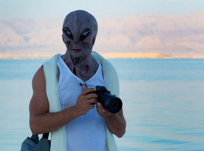 Person wearing alien mask holding camera by the ocean, symbolizing raw and emotional confessions of heavy secrets.