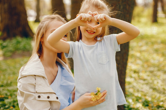 Young girl making a heart shape with hands while mother holds yellow flowers in a peaceful outdoor setting, expressing emotional confessions.