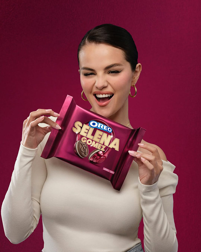 Woman holding Oreo Selena Gomez flavored cookies package with a playful expression on a solid burgundy background. Woman holding Oreo Selena Gomez flavored cookies package with a playful expression on a solid burgundy background.
