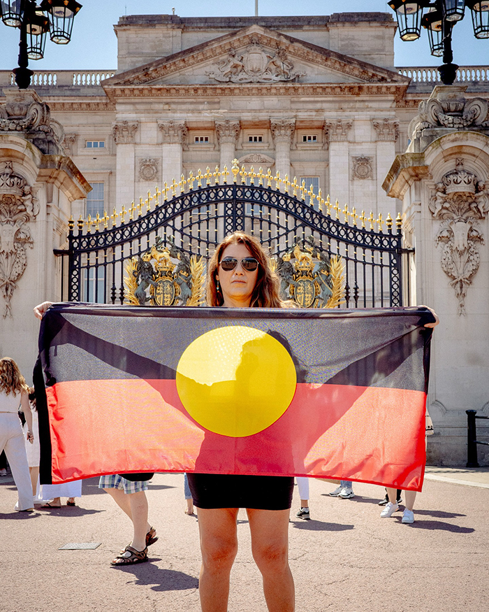Aboriginal artist holding flag outside Buckingham Palace, sparking debate related to King Charles controversy. Aboriginal artist holding flag outside Buckingham Palace, sparking debate related to King Charles controversy.