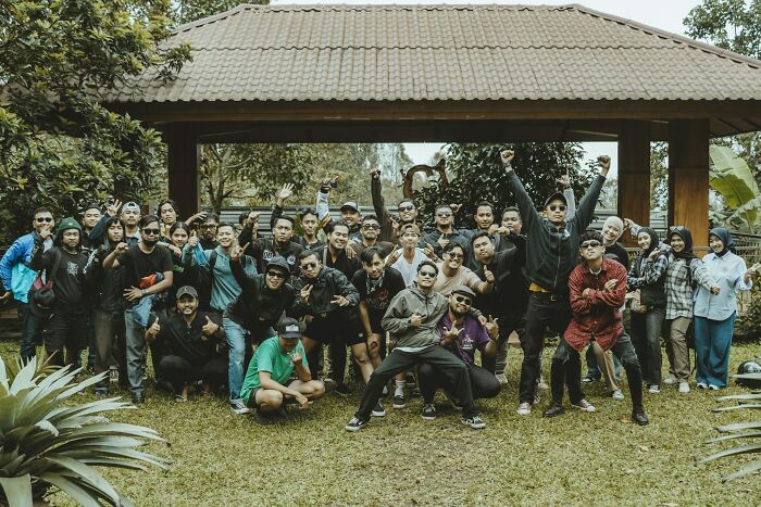 A large group of people outdoors, posing energetically in front of a wooden shelter, showcasing diverse expressions and gestures.