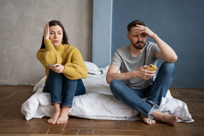 Frustrated man and woman sit apart on bed after disagreement about yawning, showing tension in bedroom setting.