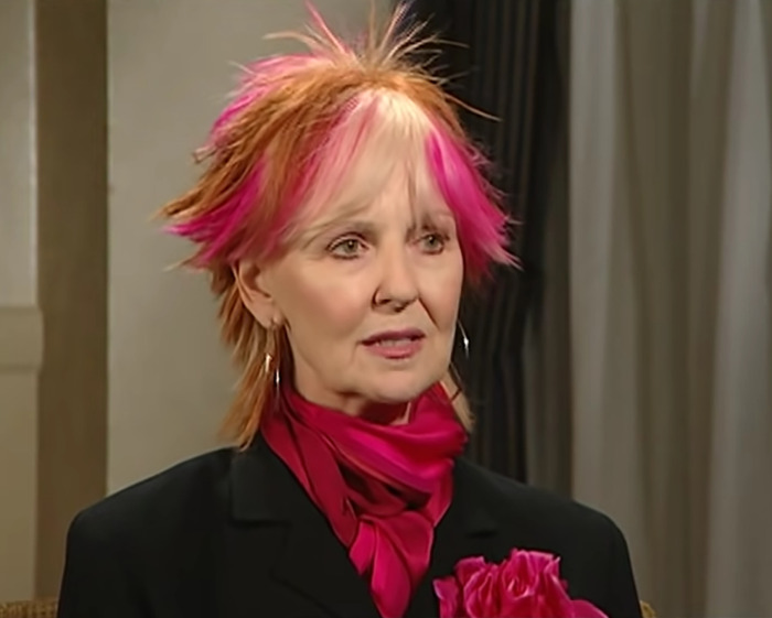 Sitcom legend with vibrant pink and orange hair, wearing black suit and matching pink scarf and flower during interview.
