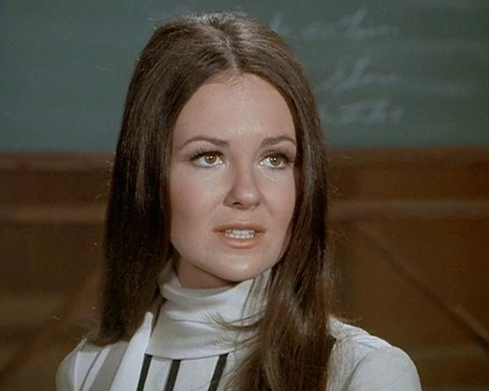 Young woman with long brown hair and a white turtleneck top in a classic sitcom scene indoors