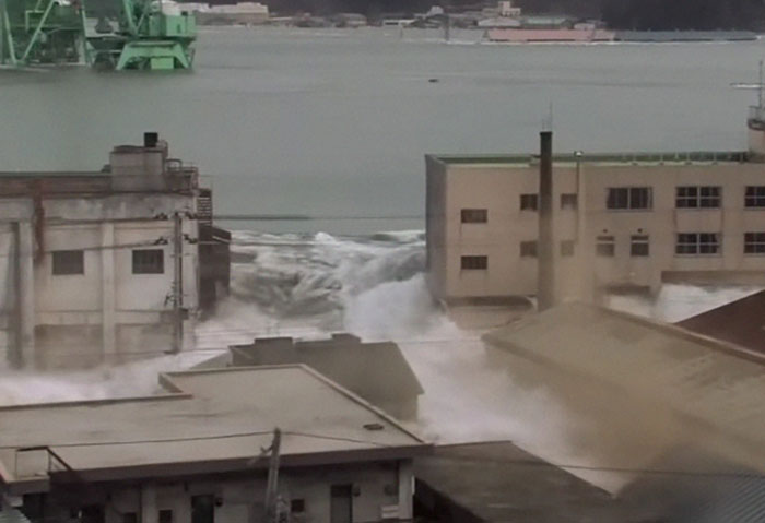 Massive wave crashing into buildings by the water, capturing one of the most shocking moments on live television.