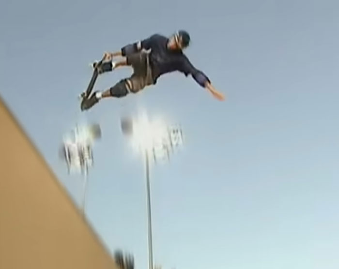 Skateboarder performing a high air trick at a skate park under bright stadium lights during an outdoor session.