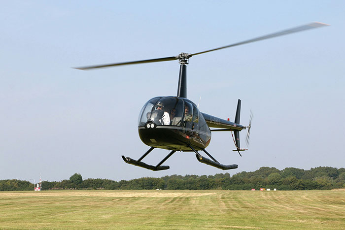 Black helicopter flying low over open grassy field with trees in the background during clear day.