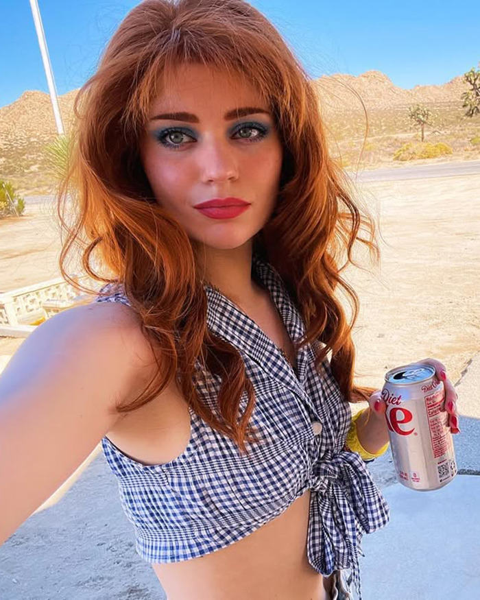 Young woman with red hair and bold makeup holds a soda can outdoors, highlighting child star's unrecognizable transformation.