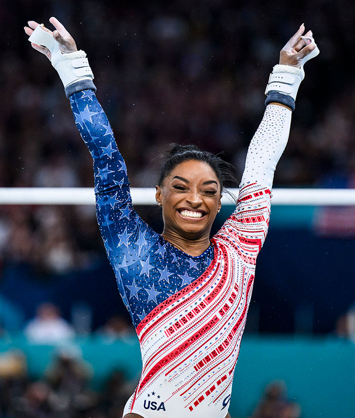 Simone Biles in USA gymnastics uniform with arms raised, highlighting her criticism of Kylie Jenner's clothing line jacket fit.