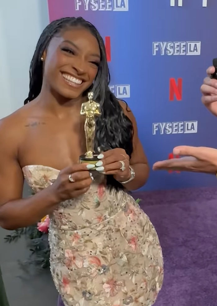 Simone Biles smiling, holding an award, wearing a floral strapless dress at a Netflix FYSEE LA event.
