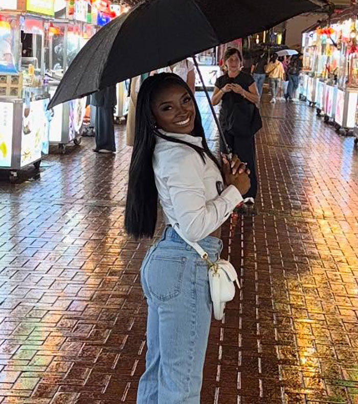 Woman smiling under umbrella on rainy street, referencing Simone Biles slamming Kylie Jenner over ill-fitting jacket.