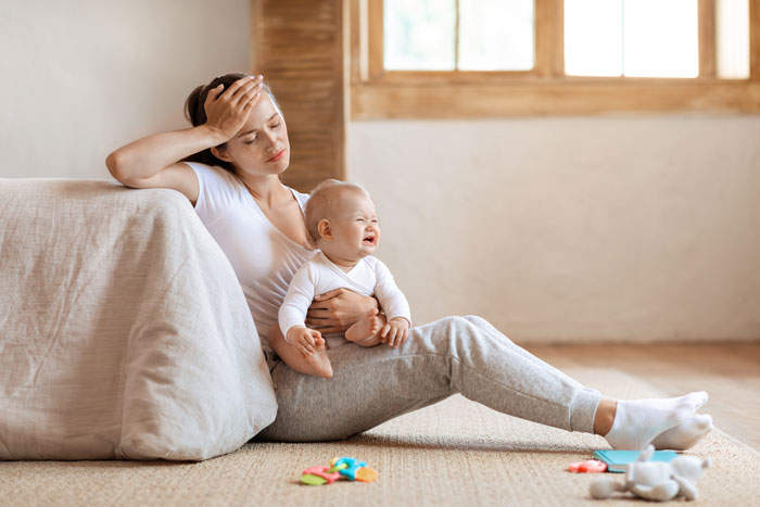Woman looking tired while sitting on the floor holding a crying baby, highlighting babysitting challenges during family visit.