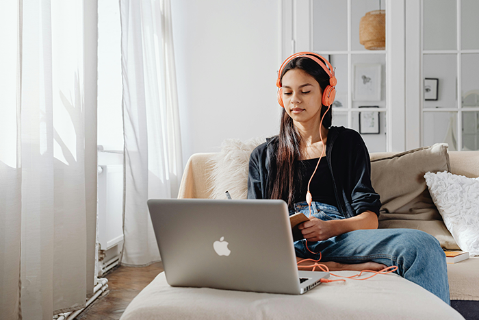 Teen sister using laptop and headphones on couch during senior year while preparing to sibling babysit at home.