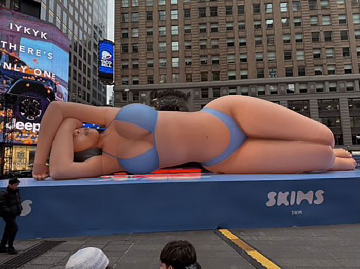 Giant billboard of Bianca Censori in a daring bodysuit with hair extensions, prompting Kim K clone comments in a city square.