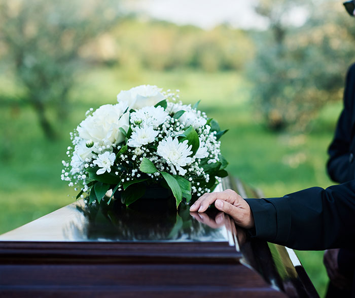 Person in dark clothing touching a casket with white flowers outdoors, related to kids' lies that got way too real.