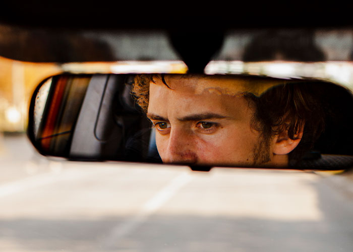 Young man reflected in car rearview mirror, focused and contemplative, symbolizing moving away and changing identity.