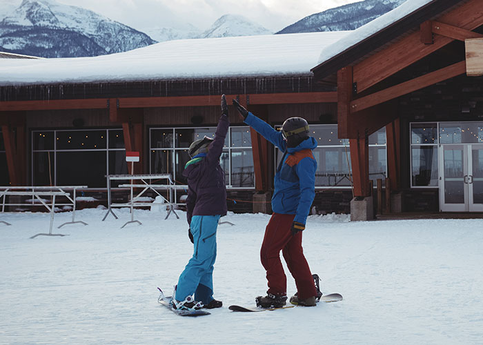 Two snowboarders in winter gear high-fiving outside a snowy lodge surrounded by mountain scenery and fresh snow.