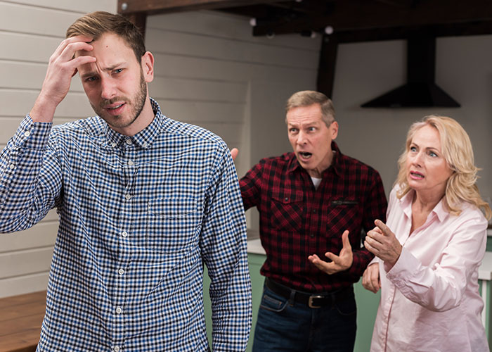 Young man looking stressed as older couple angrily confronts him indoors, depicting a life story rewrite conflict scenario.