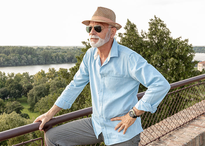Elderly man wearing hat and sunglasses, standing outdoors by railing with scenic river and trees, symbolizing life change.