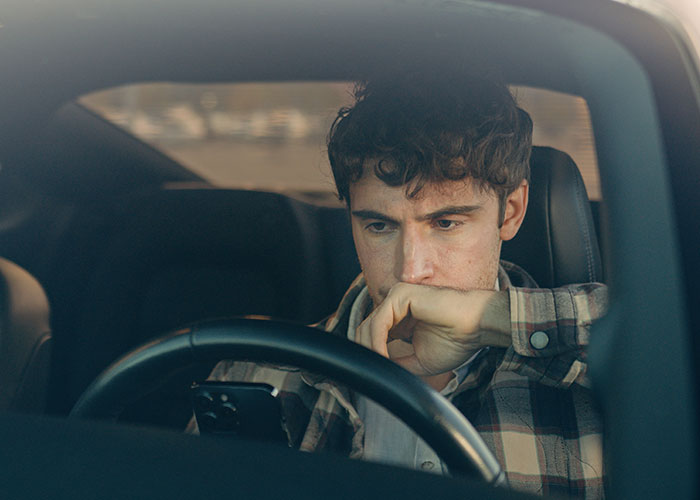 Young man inside car, looking thoughtfully at phone, possibly reflecting on moving away and rewriting his life story.