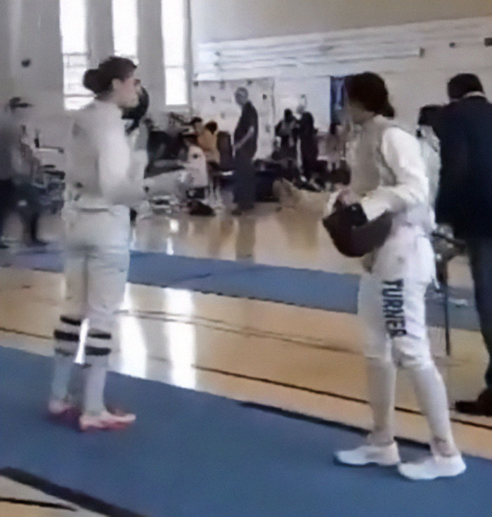 Two fencers in white gear standing and preparing to compete indoors on a blue mat in a sports setting.