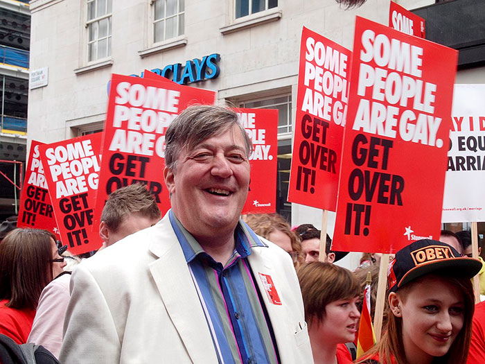 Stephen Fry at a protest holding signs supporting LGBTQ+ rights with messages about acceptance and equality.