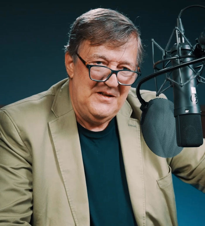 Stephen Fry speaking into a microphone, wearing glasses and a beige jacket, delivering a brutal takedown commentary.